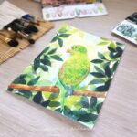 Aquarelle animale représentant une perruche verte debout sur une branche, camouflée dans les feuillages verts, en cours de réalisation par Christelle Lachambre artiste-peintre